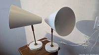 Pair of two adjustable metal cone lamps with white bases and brass stems, shown side by side on a wooden surface against a light wall.