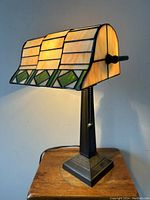 Front and side views of 16-inch stained glass bankers desk lamp lit, showing beige and green stained glass shade with geometric pattern and dark metal base with decorative embossed design and pull chain switch.