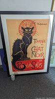 Full view of the framed "Tournee De Chat Noir" poster leaning against a wall with a soda can for scale, showing the entire black cat illustration and text.