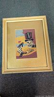 Full view of framed needlepoint depicting skull with top hat sitting at a table with pistol, cards, and drink.
