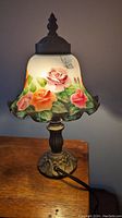 Front view of table lamp illuminated showing painted floral and butterfly design on stained glass shade, ornate metal base visible.