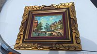 Full view of the framed oil painting showing ornate gold frame with velvet maroon mat and impressionist cityscape scene.