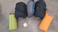 Two black sleeping bags in compression sacks, one green sleeping mat in storage sack, one orange pillow arranged on floor.