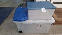 Two clear plastic Sterilite totes stacked with a blue tarp folded on top, showing condition and lot contents.