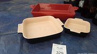 Three stoneware bakeware pieces: red long rectangular dish, large cream and black square dish, small cream and black square dish placed on blue fabric.