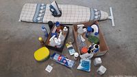 Full view of the ironing board with iron and two open boxes containing assorted cleaning liquids, sprays, and bottles, along with a jar of Vaseline and a packet of blue cleaning cloths