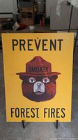 Yellow rectangular wooden sign with Smokey the Bear face and text 'Prevent Forest Fires', on a black metal display stand. Some scratches visible on the painted surface.