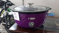 Side view of purple Bella slow cooker showing diamond textured exterior, metal Bella badge, control knob, and glass lid.