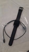 Black smart watch laid flat with attached black charging cable showing front display and strap