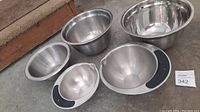 Five stainless steel mixing bowls arranged on floor displaying various sizes and styles, including two with black silicone handles and pouring spouts, identified as Hamilton Beach brand.