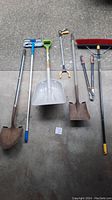Full view of yard tools laid on ground including shovels, snow shovel, grabber, pruning shears, and broom