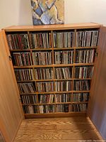 Wooden cabinet with double doors opened revealing six shelves filled with CD jewel cases.