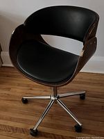 Desk chair angled left showing wood veneer shell with wear, black padded seat and backrest, and chrome base with casters