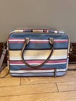 Front view of laptop bag showing horizontal multicolor stripes in blue, pink, yellow, white, and brown. Two beige handles and structured rectangular shape.