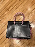 Front view of black leather purse with pink handles and pink bottom trim, showing design details on hardwood floor.