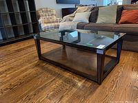 Glass top coffee table with dark wood frame and lower shelf, viewed from front left corner angle.
