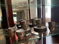 Photo showing full set on glass shelf including cups, side plates stacked, saucers stacked, creamer, and sugar bowl.