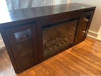 Front view of dark wood cabinet with electric fireplace insert center with glass-front storage doors on either side.