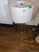 Photo showing white cloth drum lampshade and detailed metal circular loop patterned base, lamp plugged in and cords visible on wooden floor