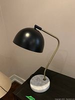 Photo showing desk lamp with black metal shade, brass-colored curved arm, and round white marble base on a black surface against beige wall.