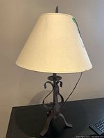 Front view of the table lamp showing the cloth lampshade and iron scroll base on a dark surface against a beige wall.