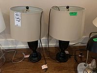 Two metal base table lamps with beige cloth shades standing side by side on a wooden floor against a white wall. Both lamps measure approximately 24 inches tall.