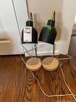 Photo showing the two metal desk lamps side by side with black shades and light wood circular bases on a wooden floor near a white door.