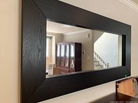 Front view of large rectangular wall mirror with thick dark wood frame reflecting furniture and stairway.