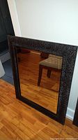 Framed beveled mirror leaning against wall on hardwood floor showing full mirror and frame with chair reflection.