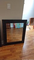 Framed bevelled mirror with textured dark frame leaning against a beige wall showing wood floor reflection