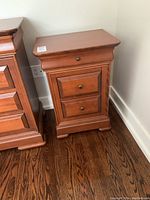 Front and left side view of the reddish-brown wooden night stand with three drawers and brass-colored round knobs, showing decorative trim and block feet base.