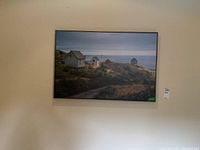 Frontal view of the large framed canvas print depicting multiple small coastal houses by the ocean, mounted on a beige wall.