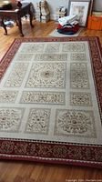 Full view of rectangular beige and red oriental style area rug with intricate geometric and floral designs in framed sections, laid out on hardwood floor with furniture and other household items around.