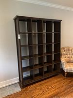 Front view of wood shelving unit showing 20 square compartments, dark wood finish, placed near wall and adjacent upholstered chair.