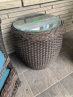 Close angle of round wicker side table with glass top in outdoor setting adjacent to chair.