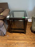 Front-facing image of a wooden side table with a clear glass top showing the overall shape, dark wood frame, and lower wood shelf, surrounded by upholstered furniture.