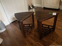 Two wooden triangular tables placed on wood floor near white doors, showing front and side angles with slatted top and bottom shelves.
