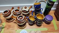 Five brown French onion soup bowls with lids, six blue and mustard yellow teacups stacked, two modern thermos water bottles, small ceramic container with purple lid on wooden table