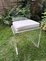 Front-left angle view of transparent lucite chair with white cushion placed outdoors on grass, surrounded by plants