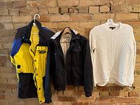 Overview of the three clothing items hung on hangers against a rustic brick wall. Left: yellow and black with blue windbreaker, center: black coat with TNA label, right: white knit sweater.