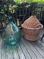 Outdoor photo showing the large green/blue glass demijohn next to the woven wicker basket with a lid, placed on a wooden deck with greenery in the background.