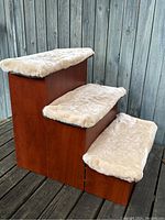 Front angle photo of three-step reddish-brown wooden dog stairs with beige faux fur pads covering each step. Shows the size and texture of the stairs and pads.