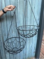 Photo showing two metal hanging basket frames, one held by hand and the other hanging, against a wooden fence background.