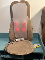Front view of brown and beige massage cushion with red glowing massage nodes and attached wired remote resting on a blue surface.