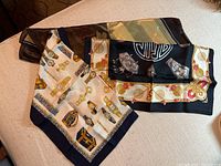 Three scarves laid out: two small square scarves (one white with watch designs, one black with floral motifs) and one larger black scarf with ornate patterns.