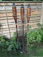 Three bamboo tiki torches standing upright in the garden next to fence and plants.