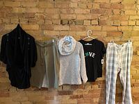 Photo of five women's clothing items on hangers against brick wall: black cardigan, olive pants, grey sweater, black graphic t-shirt, checkered pants