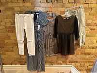 Full view of three women’s dresses and two pairs of pants hanging against a brick wall, showing styles, colors, and conditions