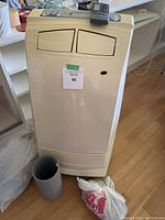 Front view of beige EnStar mobile air conditioner with two front vents, a grey cylindrical duct piece, and a plastic bag on floor next to it.