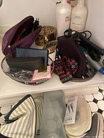 Overview of bathroom self care items including Aveda shampoo, AG conditioner, cosmetic bags, hair irons, Revlon hair dryer and silver plate vanity set on bathroom counter.